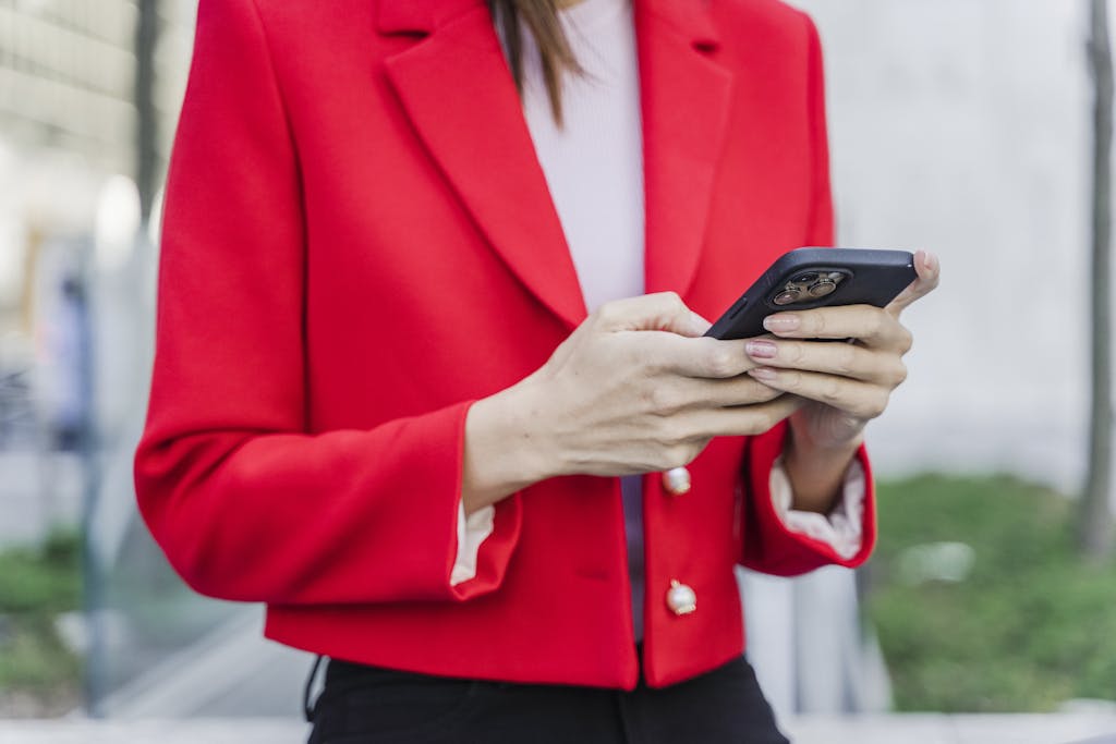 Close-up of a woman in a vibrant red blazer using a smartphone outdoors during daytime.